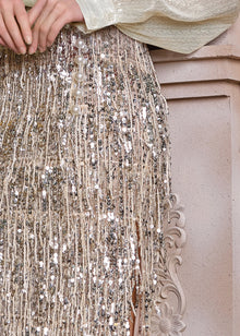 A close-up shows a hand resting on the Social Queen Sequin Fringe Side-Slit Midi Skirt, featuring fringe trim and pearl buttons, against an ornate, light-toned background.