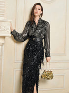 A woman in a metallic blouse and Social Queen's Sequin Fringe Side-Slit Midi Skirt holds a gold handbag, standing indoors against a cream-colored wall.