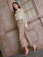 A woman poses indoors in high heels, wearing the Social Queen Sequin Fringe Side-Slit Midi Skirt and a shiny, long-sleeved blouse beside a decorative wall and window.