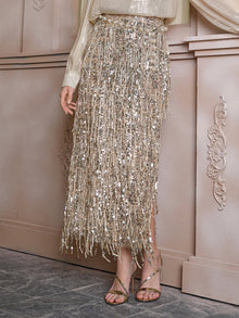 A person wearing the Social Queen Sequin Fringe Side-Slit Midi Skirt, a shimmery long-sleeve top, and strappy gold heels stands against a decorative beige wall.
