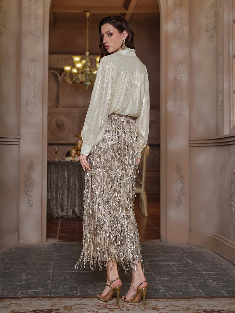 A woman stands indoors wearing the Social Queen Sequin Fringe Side-Slit Midi Skirt, paired with a shiny long-sleeved blouse and high-heeled sandals, looking over her shoulder.