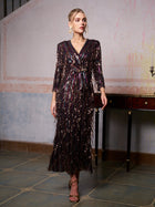 A woman stands indoors in the Social Queen Sequin Fringe Slit Evening Gown, styled with metallic heels and large earrings, posed beside a dark wooden table against a textured wall.