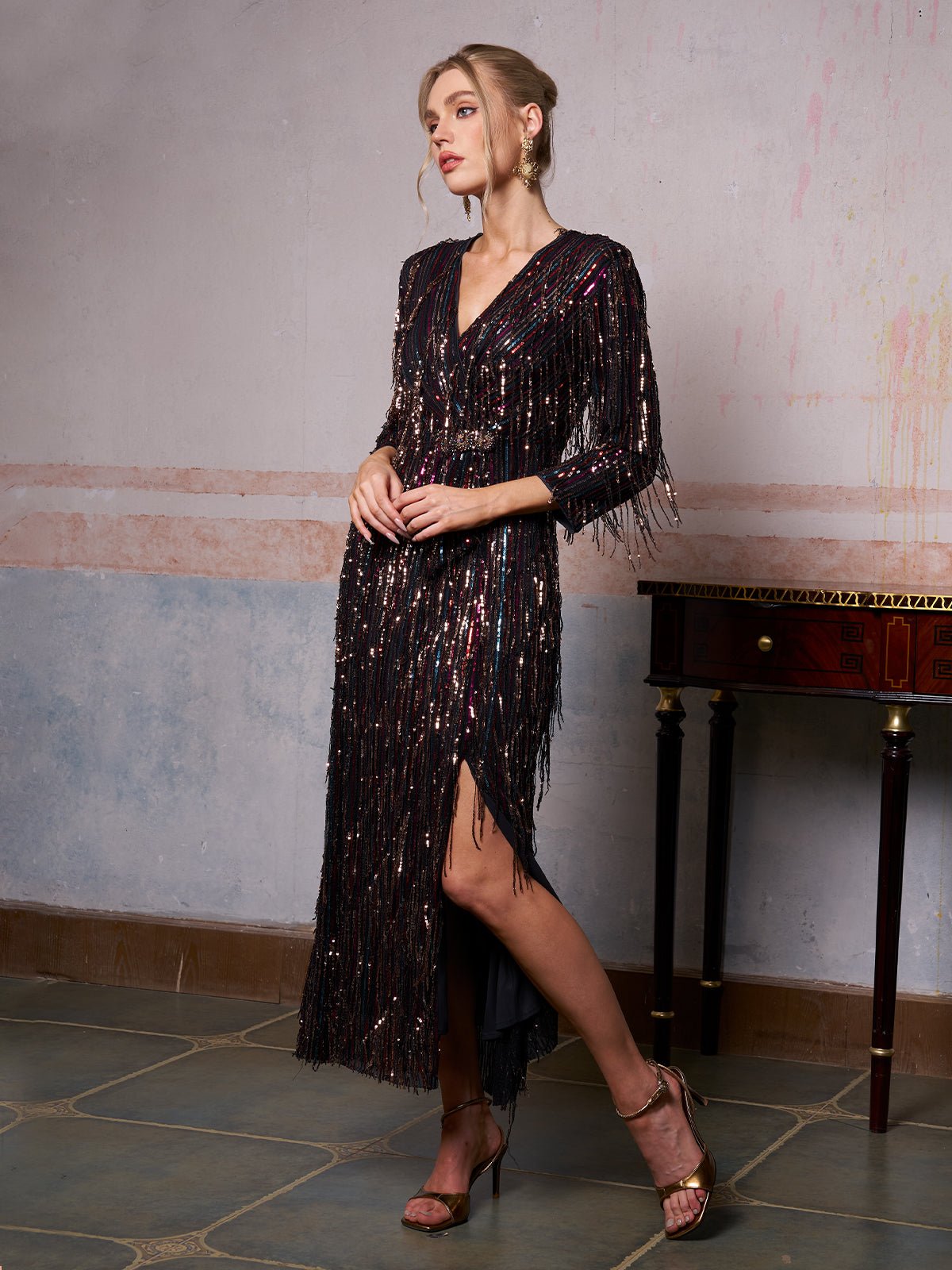 A woman wearing the Social Queen Sequin Fringe Slit Evening Gown stands indoors beside a small wooden table, looking to the side.