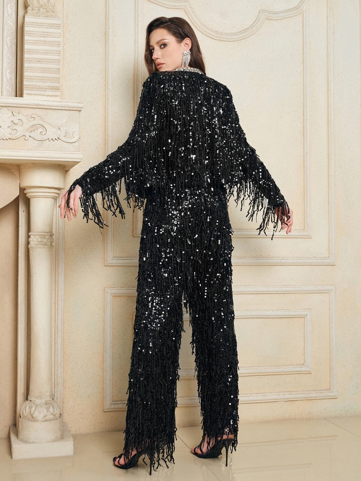A woman indoors, facing away from the camera in front of an ornate cream wall, wears Social Queen’s Sequin Fringe Wide-Leg Pants and black heels.