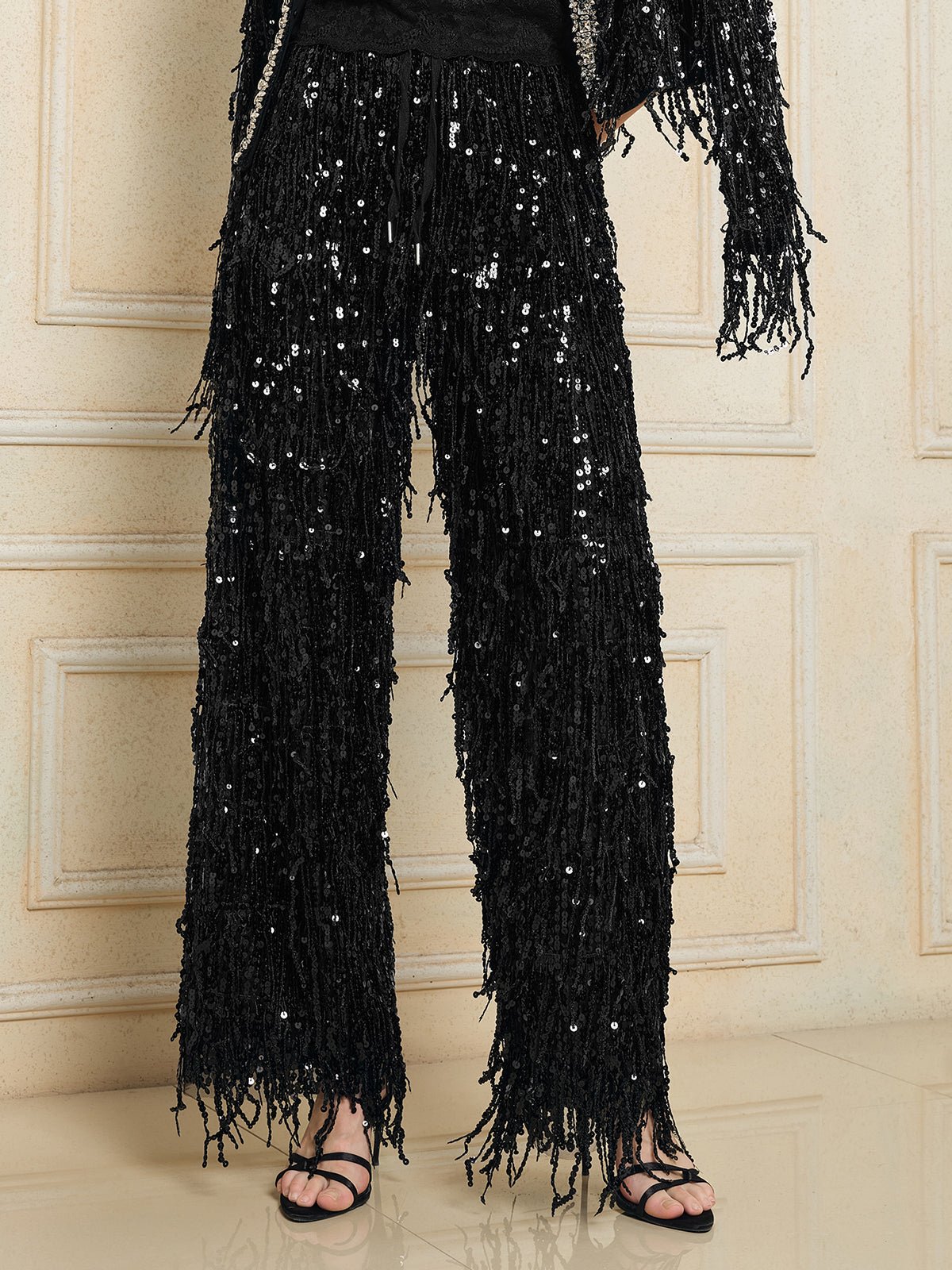 Wearing Social Queen’s Sequin Fringe Wide-Leg Pants with black strappy sandals, this standout look pops against a beige paneled wall.