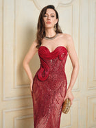 A woman stands against a cream wall, holding a gold clutch and wearing drop earrings and a necklace, dressed in Social Queen’s Sequin & Hand-Beaded 3D Pleated Tulle Gown with a strapless heart neckline and swirl designs.