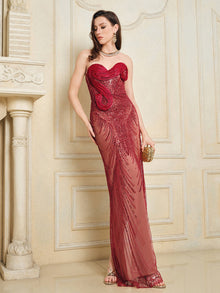 A woman in Social Queen’s Sequin & Hand-Beaded 3D Pleated Tulle Gown, a floor-length red strapless piece, holds a gold clutch while standing before an ornate cream-colored wall.