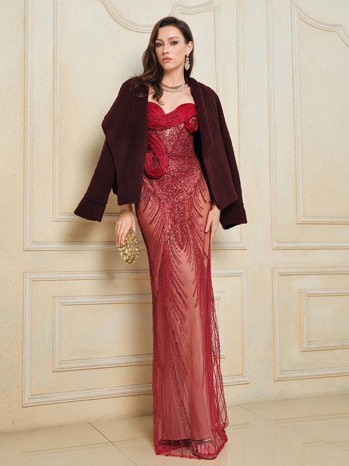 A woman wears the Social Queen Sequin & Hand-Beaded 3D Pleated Tulle Gown—a long, sheer red dress with intricate details—styled with a dark jacket and gold clutch, standing by an ornate wall.