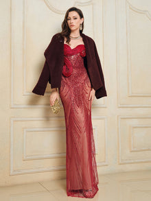 A woman wears the Social Queen Sequin & Hand-Beaded 3D Pleated Tulle Gown—a long, sheer red dress with intricate details—styled with a dark jacket and gold clutch, standing by an ornate wall.