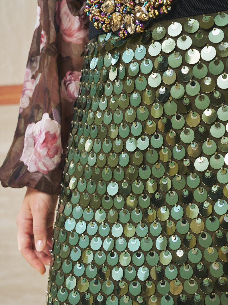 A person wearing the Social Queen Sequin Layered Slit Skirt, featuring large green metallic sequins and a jeweled waist belt, showcases stunning layered texture with sheer floral sleeves.