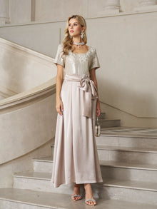 A woman stands on a staircase in the Social Queen Sequin & Satin Bow-Waist Dress, featuring a sparkling bodice and long satin skirt. She accessorizes with statement jewelry and a small handbag.