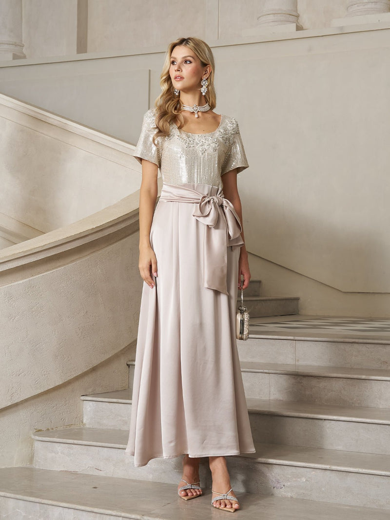 A woman stands on a staircase in the Social Queen Sequin & Satin Bow-Waist Dress, featuring a sparkling bodice and long satin skirt. She accessorizes with statement jewelry and a small handbag.
