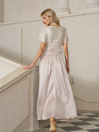 A woman in the Social Queen Sequin & Satin Bow-Waist Dress, featuring a shimmering silver sequin bodice and light pink satin skirt, stands on a staircase facing away, holding a small purse.