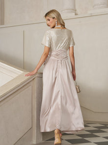 A woman in the Social Queen Sequin & Satin Bow-Waist Dress, featuring a shimmering silver sequin bodice and light pink satin skirt, stands on a staircase facing away, holding a small purse.