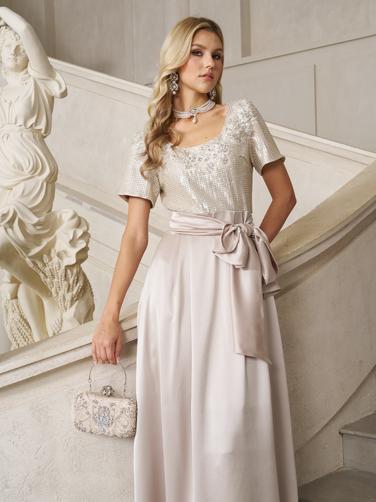 A woman in the Social Queen Sequin & Satin Bow-Waist Dress stands by a staircase, holding a decorative handbag, with a white statue in the background.
