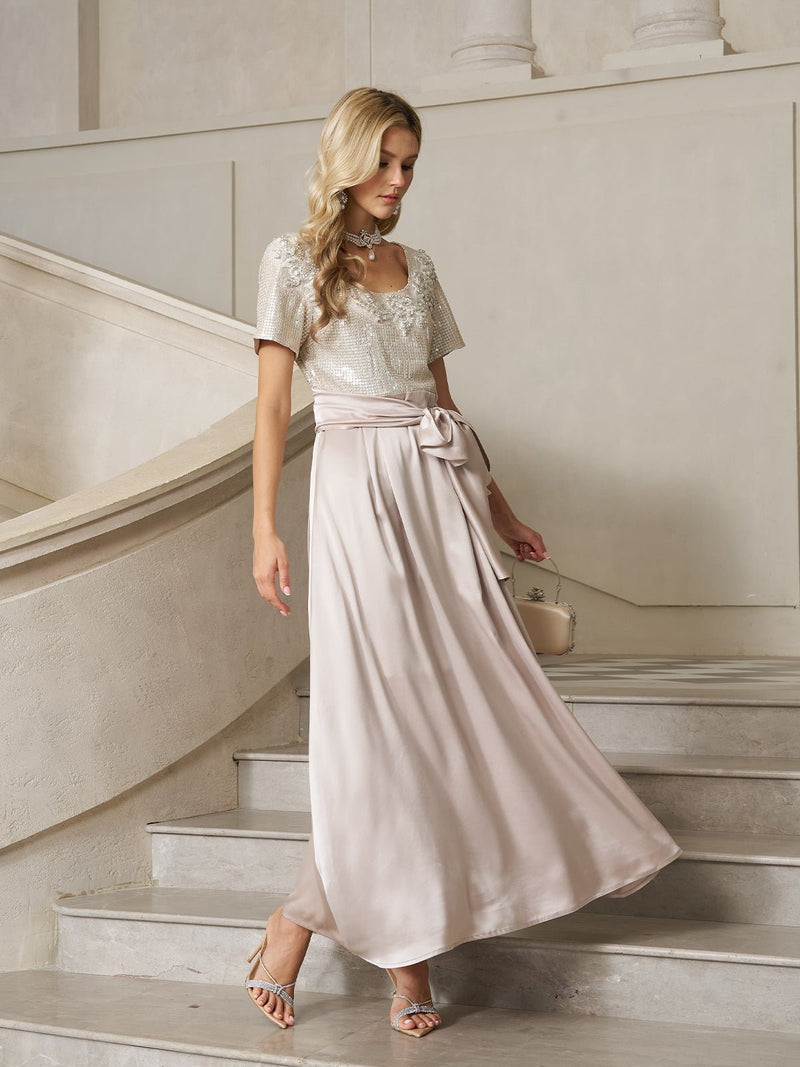 A woman in the Social Queen Sequin & Satin Bow-Waist Dress, a floor-length light pink gown with a sequin-embellished bodice and short sleeves, walks down a stone staircase holding a small handbag.