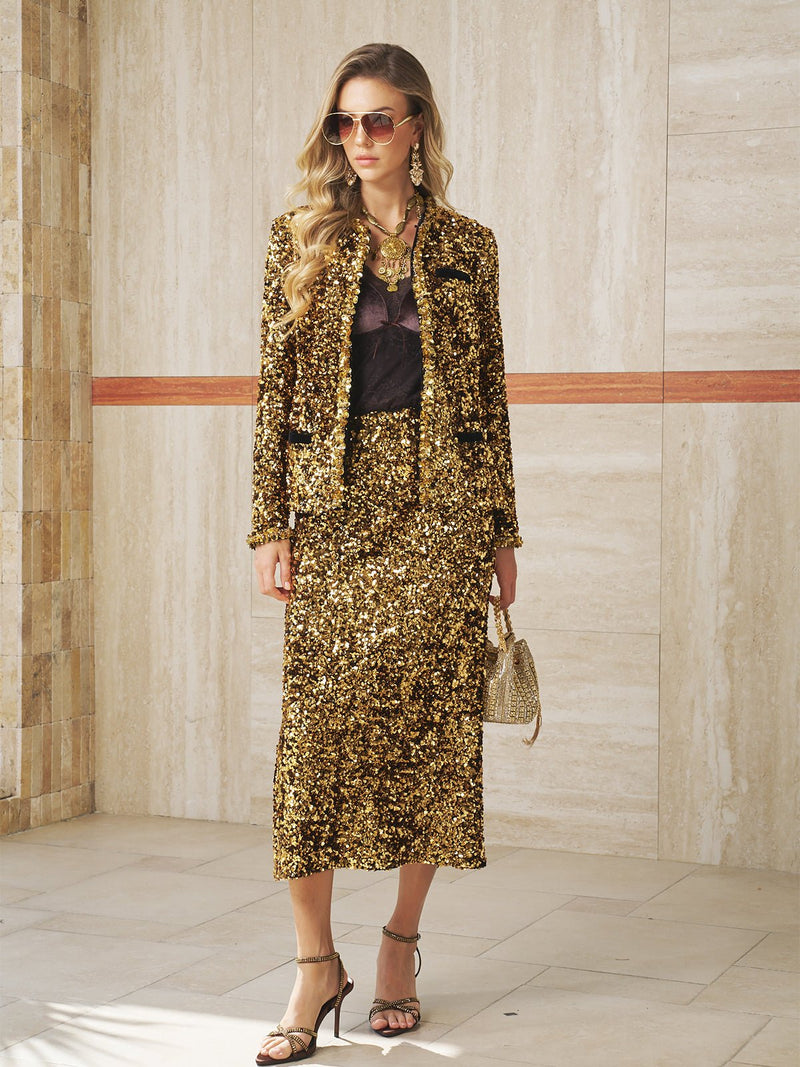 A woman stands indoors in Social Queen’s Sequin Velvet Skirt paired with a golden sequin jacket, black top, sunglasses, gold necklace, heels, and a small gold handbag—an ideal look for festive evenings.