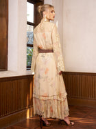 A woman stands indoors wearing the Silk Floral & Bird Print Lace Dress by Social Queen, styled with a brown belt and heeled sandals—an elegant option for a wedding guest.