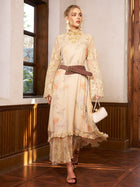A woman stands indoors on a wooden floor near a window, wearing the Social Queen Silk Floral & Bird Print Lace Dress with a brown belt and holding a small purse—an elegant option for wedding guests.