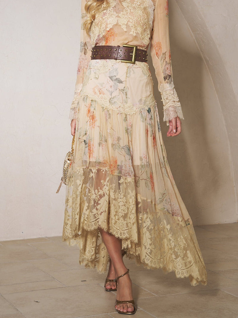A person in a Social Queen Silk Floral Lace Asymmetric Skirt, styled with a brown belt, small purse, and sandals, stands on a tiled floor.