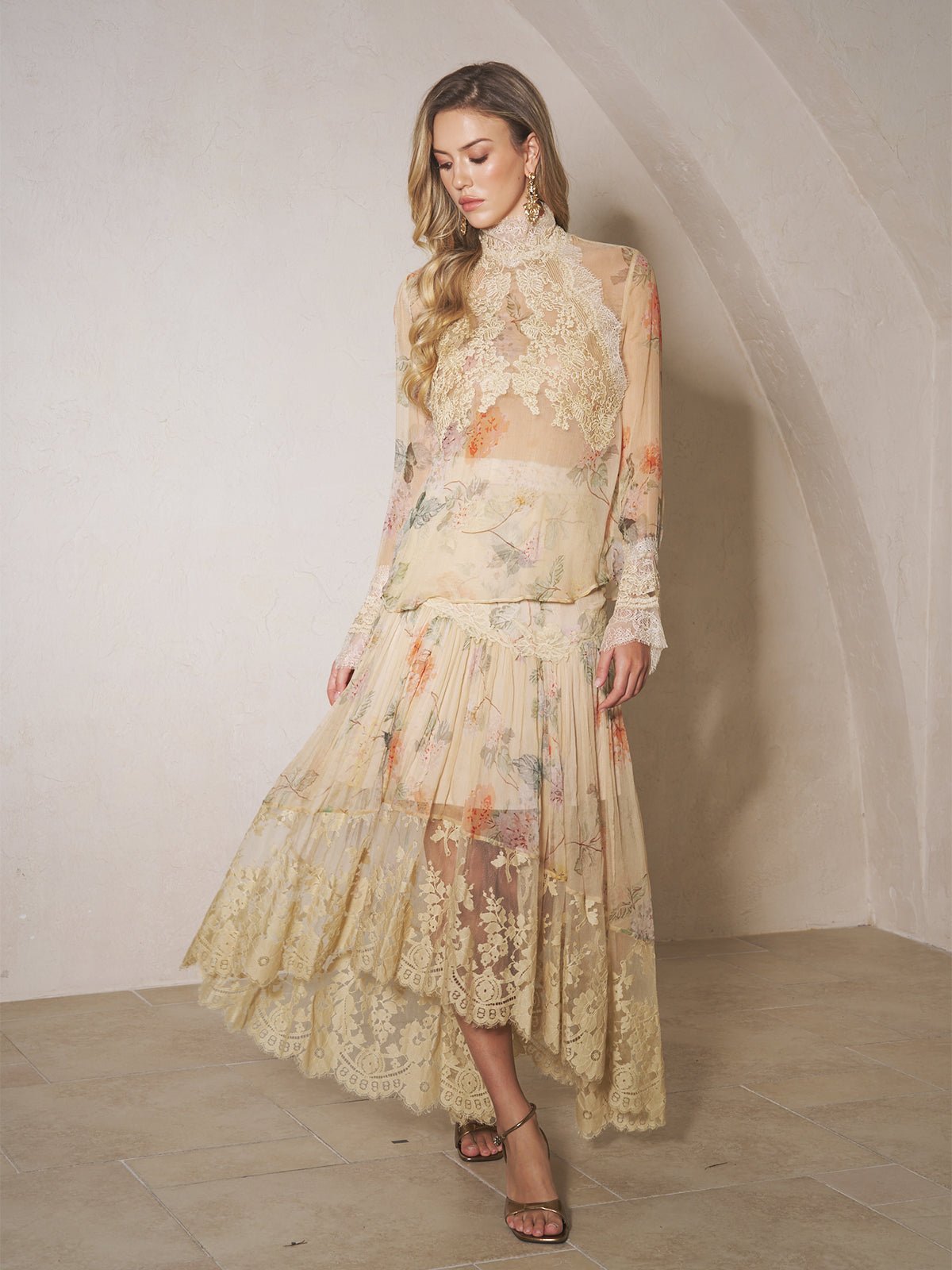 A woman stands indoors against a light-colored wall, wearing the Social Queen Silk Floral Print Lace Blouse featuring sheer panels and a high-neck design.