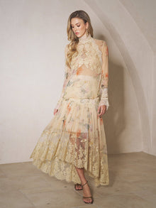 A woman stands indoors against a light-colored wall, wearing the Social Queen Silk Floral Print Lace Blouse featuring sheer panels and a high-neck design.