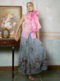 A woman wears the Social Queen Silk Organza Bow-Tie Sleeveless Top in pink and a long blue floral skirt, standing indoors beside a wooden table adorned with framed art and decor.