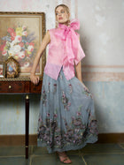 A woman wears the Social Queen Silk Organza Bow-Tie Sleeveless Top in pink and a long blue floral skirt, standing indoors beside a wooden table adorned with framed art and decor.