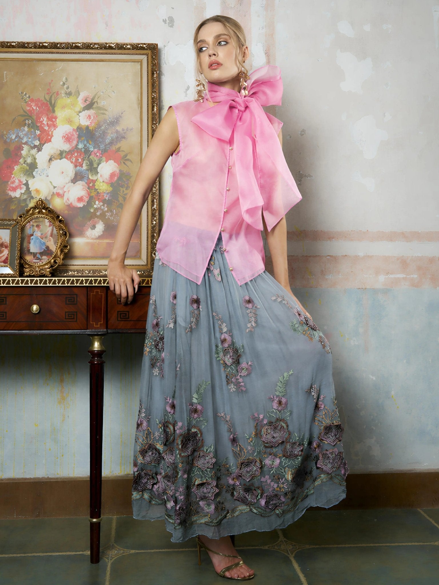 A woman wears the Social Queen Silk Organza Bow-Tie Sleeveless Top in pink and a long blue floral skirt, standing indoors beside a wooden table adorned with framed art and decor.