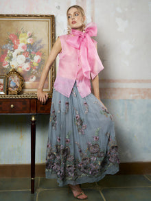 A woman wears the Social Queen Silk Organza Bow-Tie Sleeveless Top in pink and a long blue floral skirt, standing indoors beside a wooden table adorned with framed art and decor.