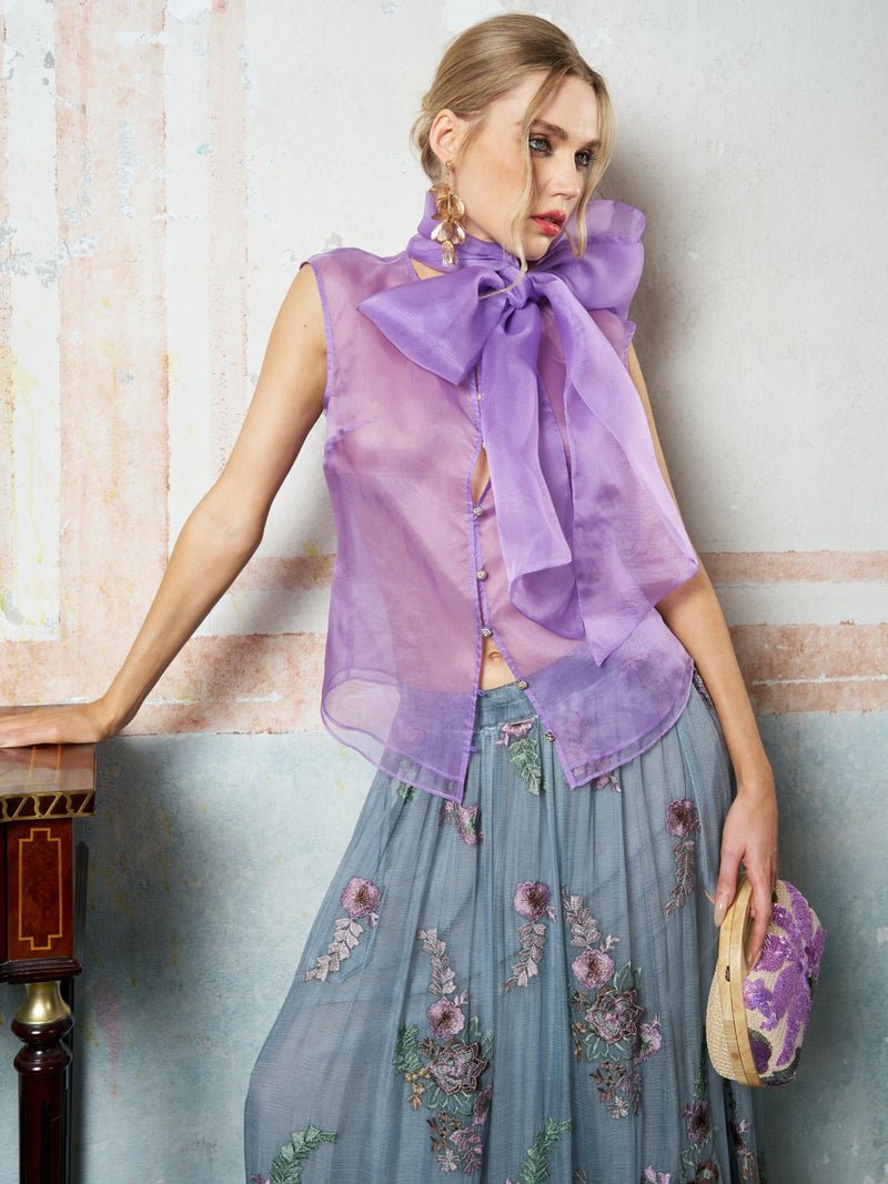A woman wears the Social Queen Silk Organza Bow-Tie Sleeveless Top in lavender with a long blue floral skirt, holding a small purple bag against a wall.