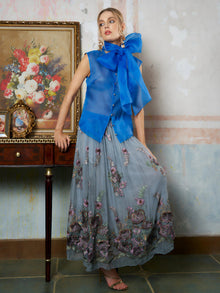 A woman wears the Social Queen Silk Organza Bow-Tie Sleeveless Top with a long floral skirt, standing by a wooden table adorned with framed art and a floral painting on the wall behind her.