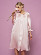 A woman in the Social Queen Silk Ruffled Shirt Sleep Dress holds a white flower near her face, posing against a solid pink background.