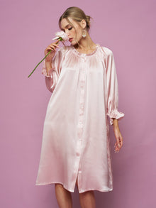 A woman in the Social Queen Silk Ruffled Shirt Sleep Dress holds a white flower near her face, posing against a solid pink background.