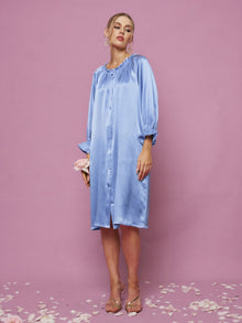 A woman stands on a pink floor with petals, wearing the Social Queen Silk Ruffled Shirt Sleep Dress in light blue satin and clear heels, against a pink background.
