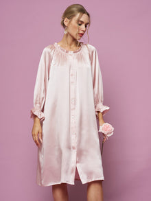 A woman in the Social Queen Silk Ruffled Shirt Sleep Dress stands against a pink background, holding a pink rose and looking down.