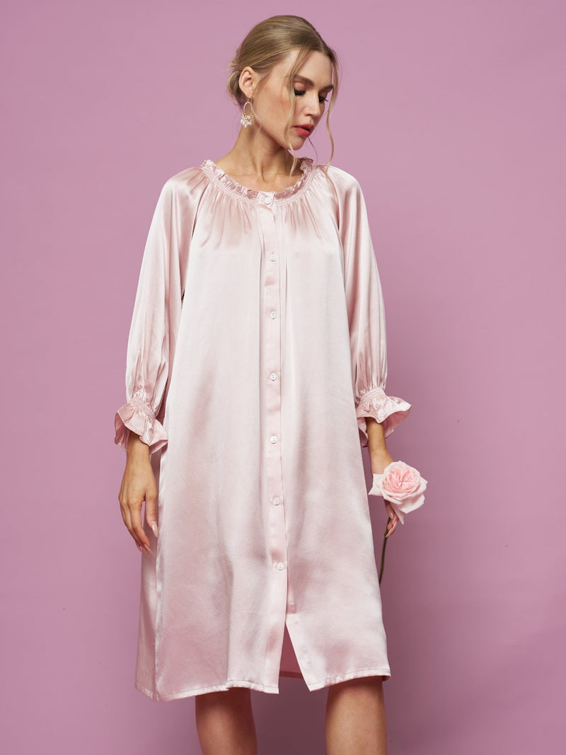 A woman in the Social Queen Silk Ruffled Shirt Sleep Dress stands against a pink background, holding a pink rose and looking down.