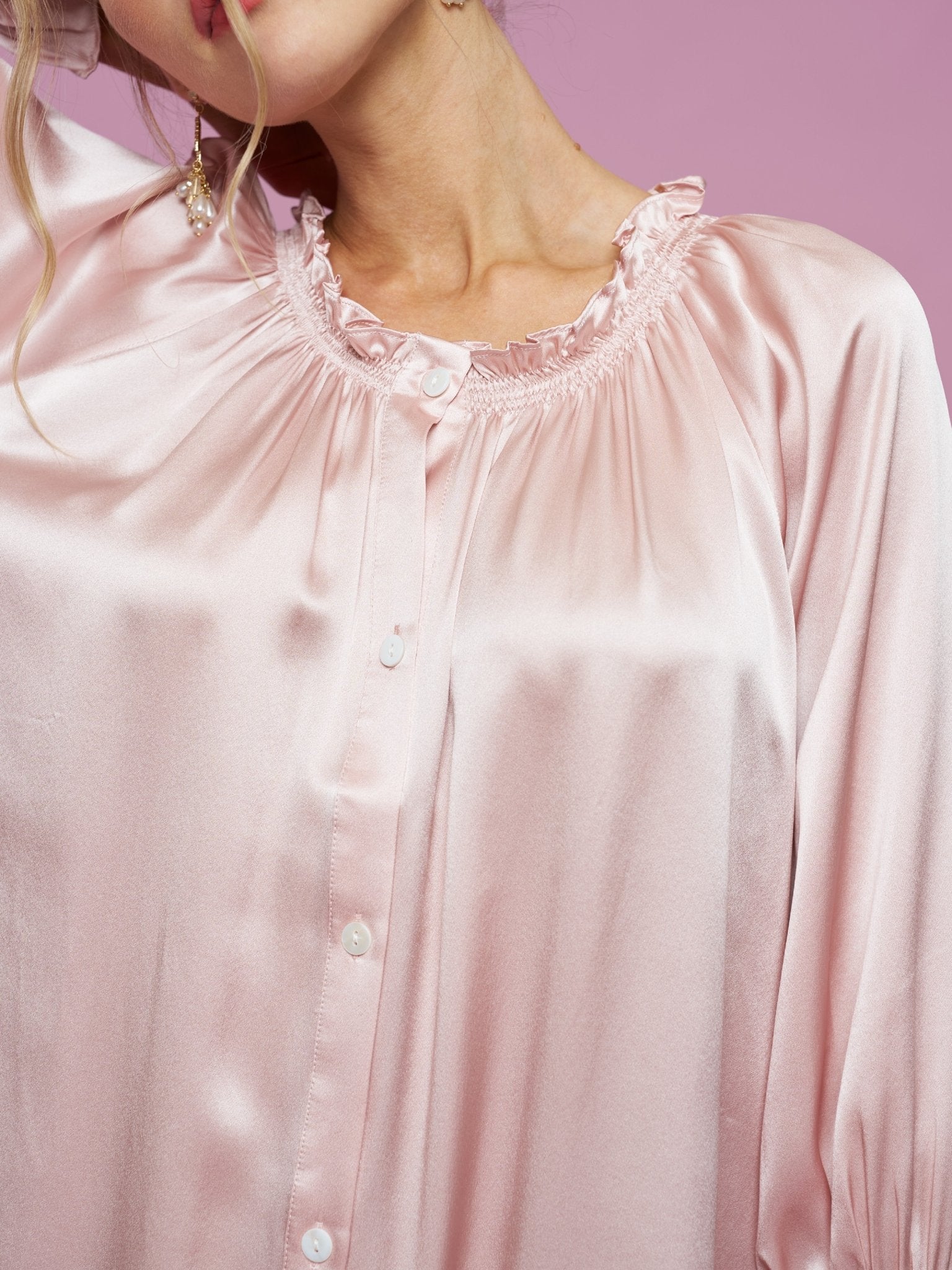 A woman wears the Social Queen Silk Ruffled Shirt Sleep Dress in shiny light pink satin with ruffle details and white buttons, set against a pink background.