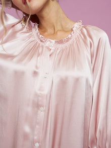 A woman wears the Social Queen Silk Ruffled Shirt Sleep Dress in shiny light pink satin with ruffle details and white buttons, set against a pink background.