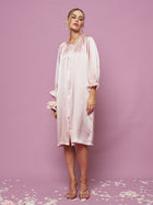 A woman stands against a pink background wearing the Social Queen Silk Ruffled Shirt Sleep Dress—a loose, knee-length silk satin dress with long sleeves and ruffle details. Rose petals are scattered on the floor.