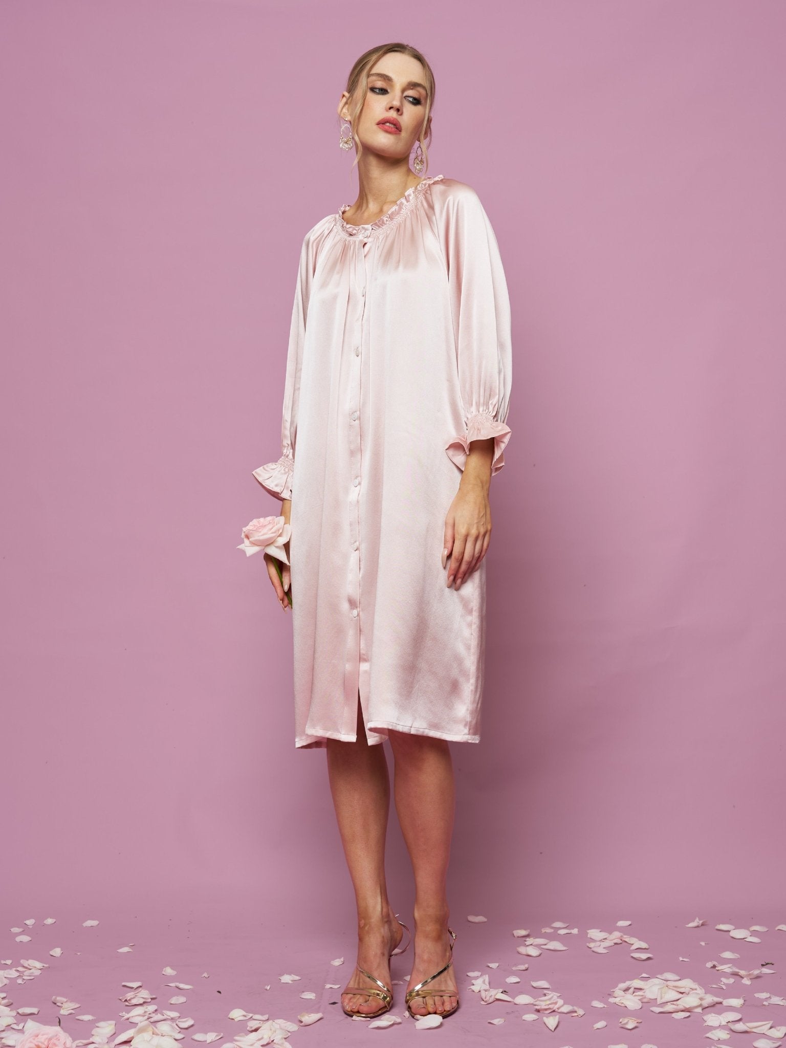 A woman stands against a pink background wearing the Social Queen Silk Ruffled Shirt Sleep Dress—a loose, knee-length silk satin dress with long sleeves and ruffle details. Rose petals are scattered on the floor.