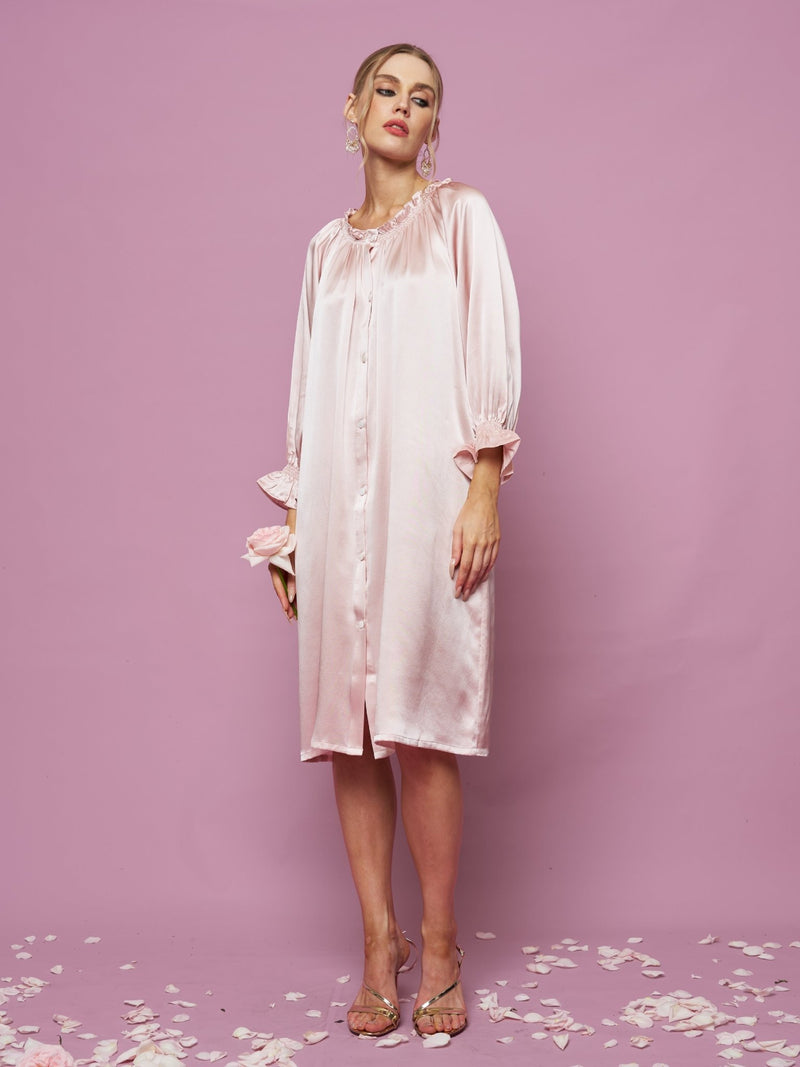 A woman stands against a pink background wearing the Social Queen Silk Ruffled Shirt Sleep Dress—a loose, knee-length silk satin dress with long sleeves and ruffle details. Rose petals are scattered on the floor.
