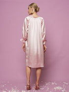 A woman stands with her back to the camera against a pink background in the Social Queen Silk Ruffled Shirt Sleep Dress, light pink and long-sleeved, with ruffle details. Flower petals are scattered on the floor.
