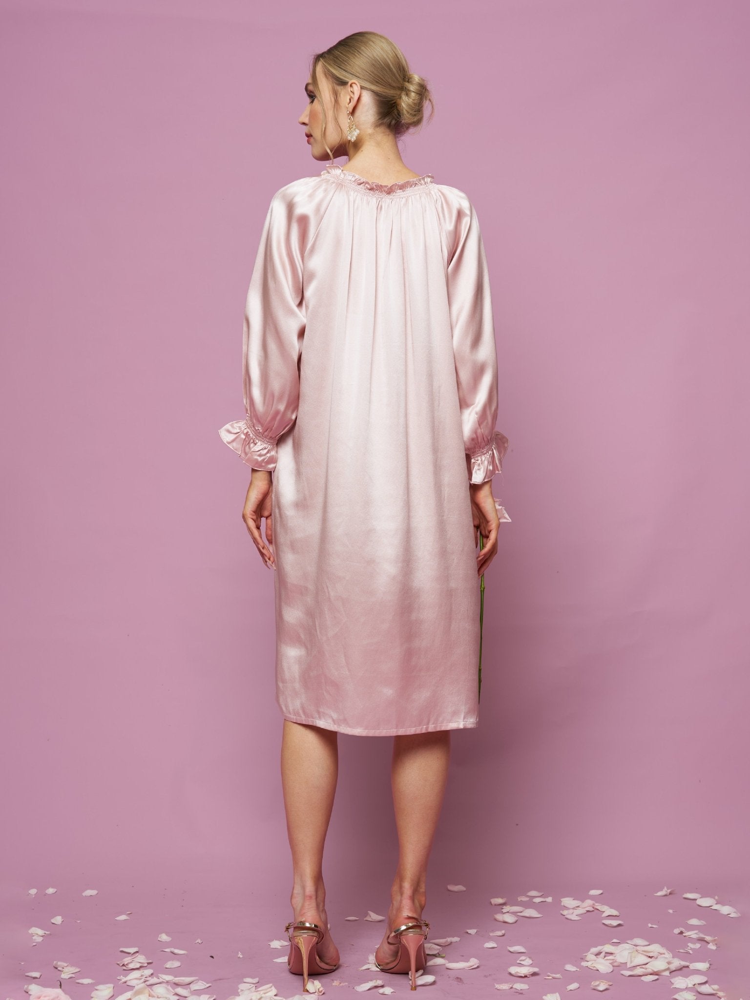 A woman stands with her back to the camera against a pink background in the Social Queen Silk Ruffled Shirt Sleep Dress, light pink and long-sleeved, with ruffle details. Flower petals are scattered on the floor.