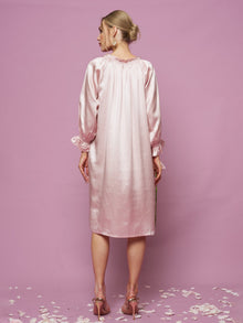 A woman stands with her back to the camera against a pink background in the Social Queen Silk Ruffled Shirt Sleep Dress, light pink and long-sleeved, with ruffle details. Flower petals are scattered on the floor.