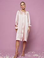 A woman poses against a pink background wearing the Silk Ruffled Shirt Sleep Dress by Social Queen. The loose, long-sleeved style drapes elegantly while flower petals are scattered around her feet.