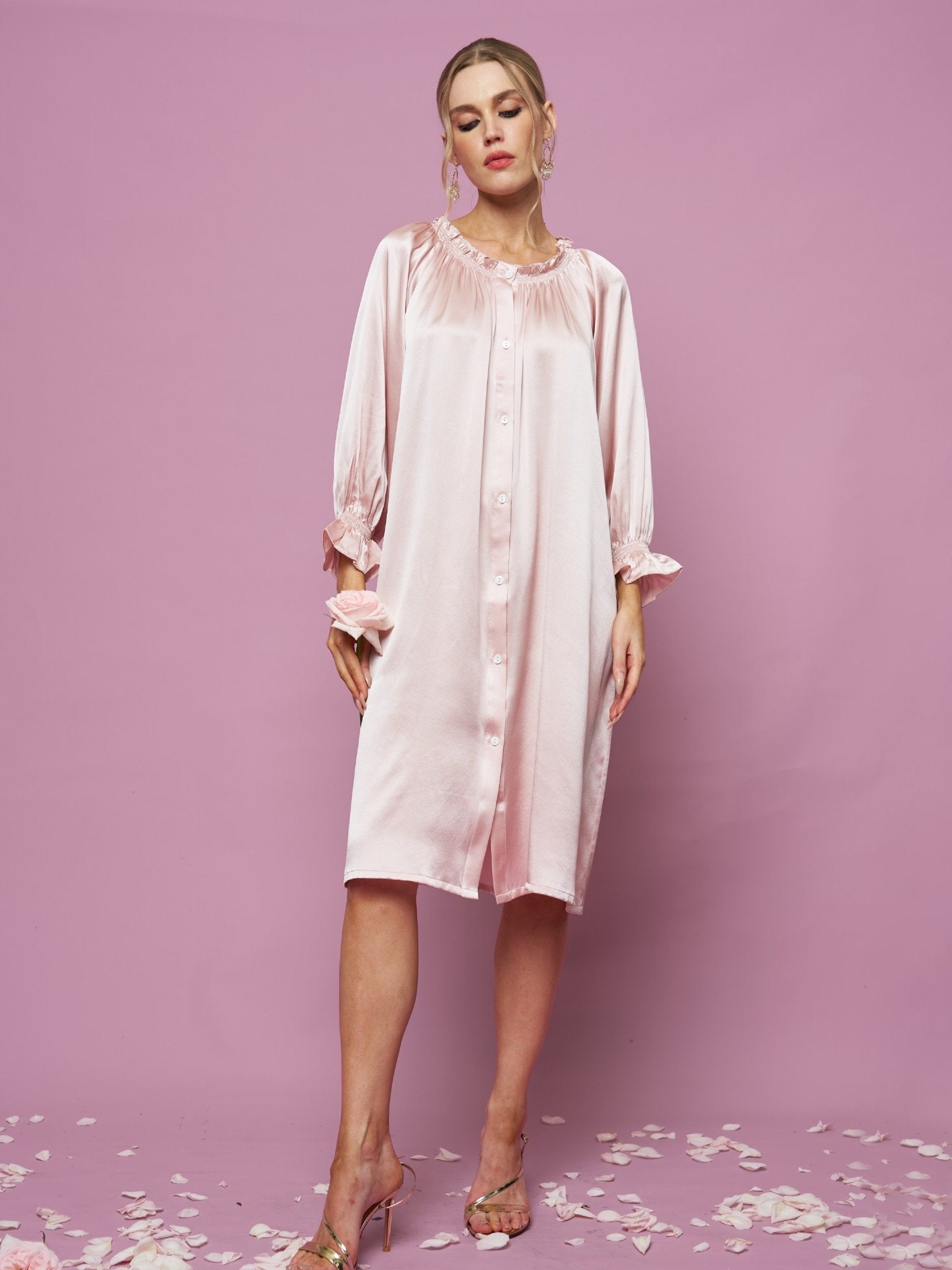 A woman poses against a pink background wearing the Silk Ruffled Shirt Sleep Dress by Social Queen. The loose, long-sleeved style drapes elegantly while flower petals are scattered around her feet.