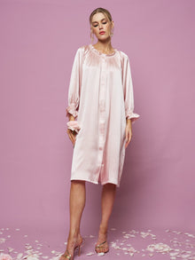 A woman poses against a pink background wearing the Silk Ruffled Shirt Sleep Dress by Social Queen. The loose, long-sleeved style drapes elegantly while flower petals are scattered around her feet.