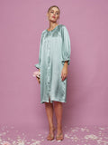 A woman wears the Social Queen Silk Ruffled Shirt Sleep Dress in light blue, standing on a pink, petal-covered floor with a pink background. She holds a pink flower and pairs the dress with clear heels.