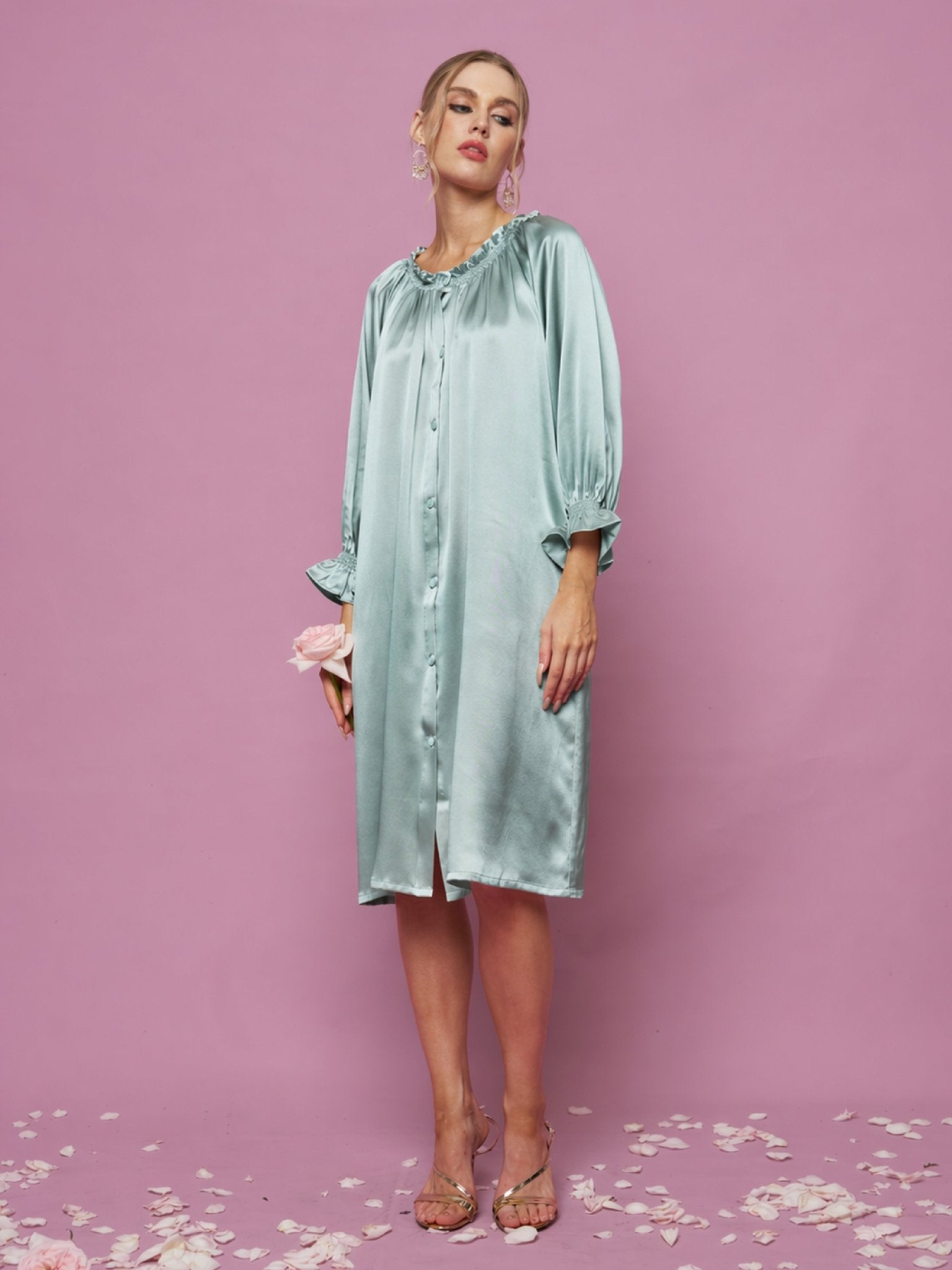A woman wears the Social Queen Silk Ruffled Shirt Sleep Dress in light blue, standing on a pink, petal-covered floor with a pink background. She holds a pink flower and pairs the dress with clear heels.